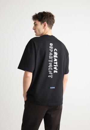 Black cotton t-shirt with short sleeves featuring vertical white text reading "CREATIVE DEPARTMENT" and a blue logo on the lower back.