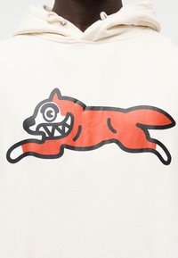 Κρεμ φούτερ με κουκούλα που απεικονίζει ένα κόκκινο σκίτσο σκύλου με υπερβολικά χαρακτηριστικά, όπως μεγάλο ανοιχτό στόμα, σε φόντο λείου υφάσματος.