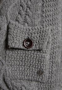 Suéter gris de punto con un patrón de cable texturizado y un bolsillo con botones. Los botones son de color marrón oscuro, lo que aporta contraste con la tela.