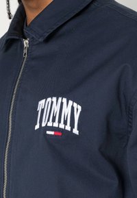 Marineblå zip-jacka med vitt broderat "TOMMY"-logotyp och röda och vita accentremsor nedanför, tillverkad av ett slätt tyg.
