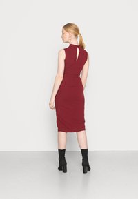 Vestido burgundy ajustado ao corpo com decote alto, recorte nas costas e comprimento até ao joelho; textura suave, cintura acentuada, combinado com botas pretas.
