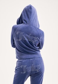 Lila velúr melegítő szett kapucnival, amelyen a hátulján és a nadrágon "Juicy" felirat csillogó strasszkövekből. Puha tapintású, testhez simuló szabás.