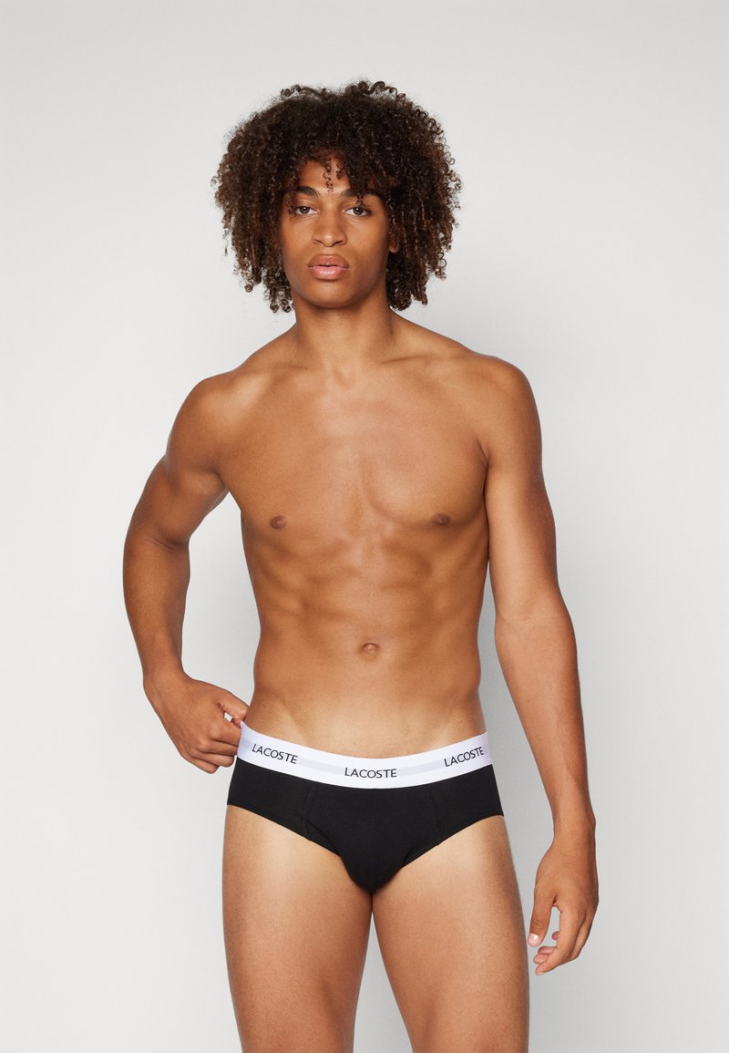 Schwarze Baumwollslips mit einem weißen Gummibund, der mit dem Schriftzug "LACOSTE" versehen ist. Das Design ist figurbetont mit einer glatten Textur.