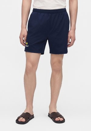 Homme portant un short bleu marine avec un petit logo jaune sur la jambe gauche, une chemise blanche et des sandales slides marron foncé, debout sur un fond uni.