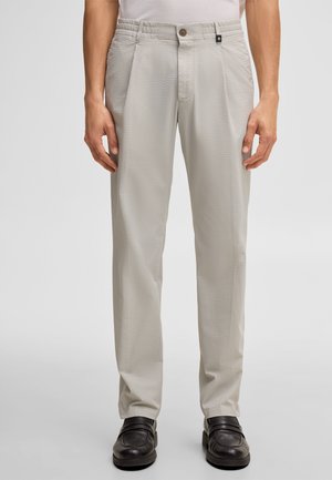 Jasnoszare spodnie o fakturze, z zakładkami, kieszeniami, zapięciem na guziki oraz czarne skórzane mokasyny na stojącej osobie.