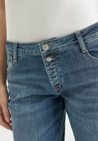 Jean en denim bleu taille haute avec une finition délavée. Dispose d'une fermeture à double bouton et de surpiqûres dorées typiques. Tissu doux et texturé.