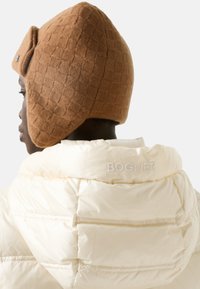 Hnědá prošívaná pletená čepice s klapkami na uši v kombinaci s krémovou péřovou bundou s vyvýšeným logem "BOGNER" na zádech.