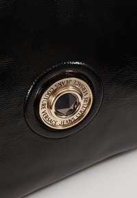 Czarna teksturowana skórzana powierzchnia z złotym okrągłym emblematem, na którym znajduje się czarny kamień w centrum oraz tekst "Versace Jeans Couture" wokół niego.