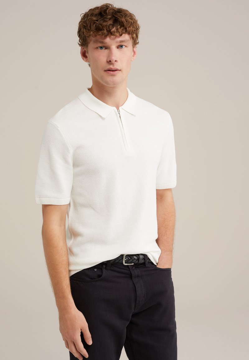 Fehér, rövid ujjú pólóing cipzáros gallérral, texturált anyaggal és bordázott mandzsettával, fekete farmerral és fekete övvel párosítva.