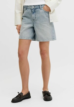 Ženska, ki nosi svetlo modre kavbojske kratke hlače, belo srajco z rumenimi črtami in črne loafer čevlje, stoji z eno roko v žepu.