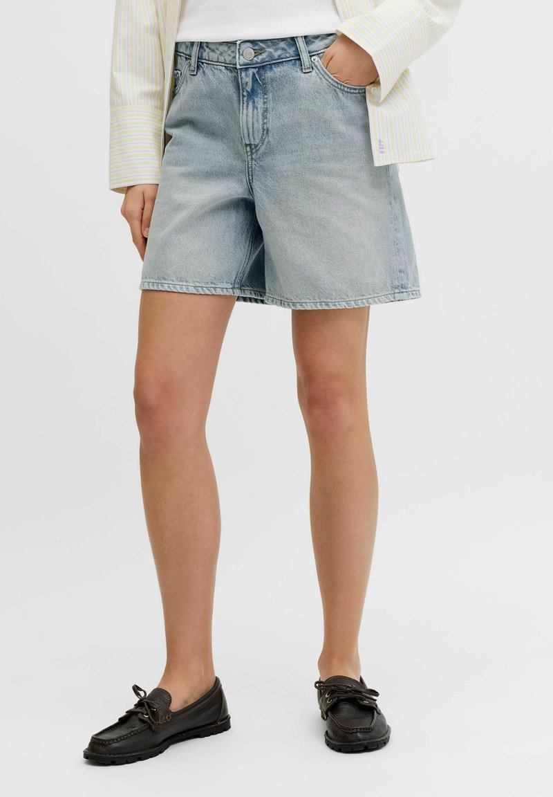 Žena v svetlomodrých riflových šortkách, bielej košeli so žltými pruhmi a čiernych mokasínach, stojaca s jednou rukou v vrecku.