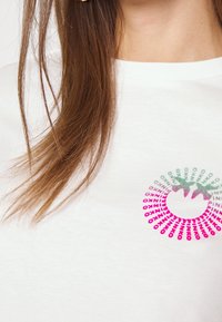 Bílý krátký rukáv tričko s kruhovým multicolor grafickým designem a textem v různých odstínech zelené a růžové, umístěným na hrudi.