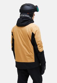 Giacca da sci con design a blocchi di colore in senape e nero, con cappuccio, superficie strutturata e dettagli di branding sul colletto.