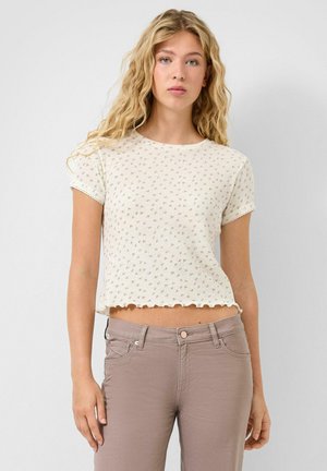 Frau mit langen, blonden lockigen Haaren, die ein weißes, gemustertes kurzärmeliges bauchfreies Oberteil und hellbraune, hochtaillierte Hosen trägt und vor einer weißen Wand steht.