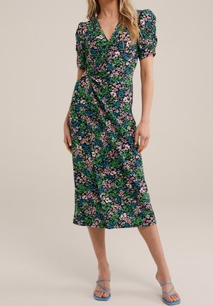 Kvinde iført en grøn floral midi kjole med pufærmer og en slå-om detalje, kombineret med blå sandaler med stropper og åben tå.