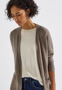 Cardigan marron clair à ouverture devant, avec des poignets côtelés et deux poches avant. Porté par-dessus un t-shirt couleur crème et un jean sombre.