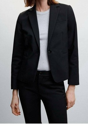 Femme portant un blazer noir cintré par-dessus un t-shirt gris clair à col rond côtelé et un pantalon noir ajusté sur un fond uni.