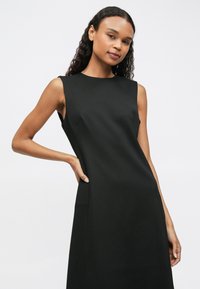 Žena s dlouhými kudrnatými vlasy, oblečená v černých bezramínkových šatech, pózující s jednou rukou na pase proti jednoduchému světlému pozadí.