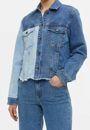 Kobieta nosząca dwukolorową kurtkę dżinsową z postrzępionym wykończeniem w połączeniu z niebieskimi, dopasowanymi jeansami z wysokim stanem, ręka w kieszeni, na białym tle.