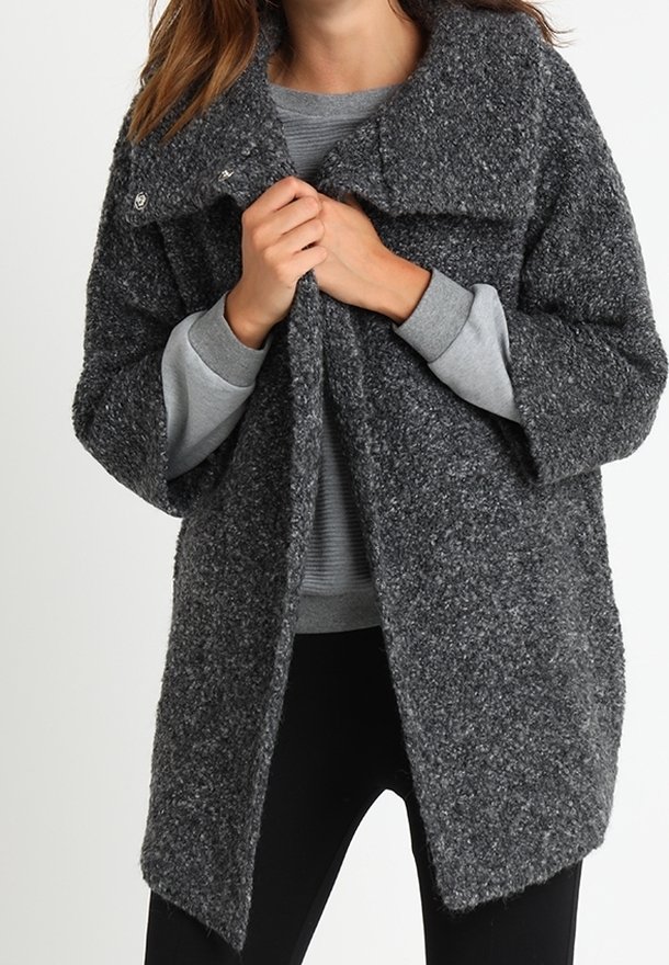 Grauer, strukturierten, übergroßer Mantel mit einem breiten Kragen und Druckknopfverschlüssen, über einem grauen Sweatshirt getragen, mit einem leicht schrägen Saum.