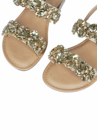 Sandali piatti beige con molteplici cinturini accentati in oro decorati con strass trasparenti e giallo pallido, dotati di una soletta in pelle liscia.