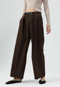 Bruine wijde broek met plooien, voorzien van een zwarte riem met een zilveren gesp. Gecombineerd met een beige aansluitend geribd topje.