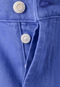 Gros plan sur un tissu en jean bleu avec deux boutons argentés portant l'inscription « JANS JEANS ESTABLISHED 1994 » sur le devant d'une veste ou d'une chemise.