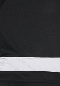 Sostenedor deportivo negro con una textura lisa y elástica, que presenta una franja horizontal blanca en la parte inferior. Diseñado para ofrecer comodidad y soporte.