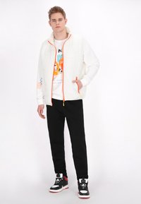 Weiß gesteppte Weste mit orangenen Reißverschlüssen, getragen über einem weißen Sweatshirt mit grafischen, bunten Texten und Designs. Schwarze Hose und hochgeschnittene Sneaker.