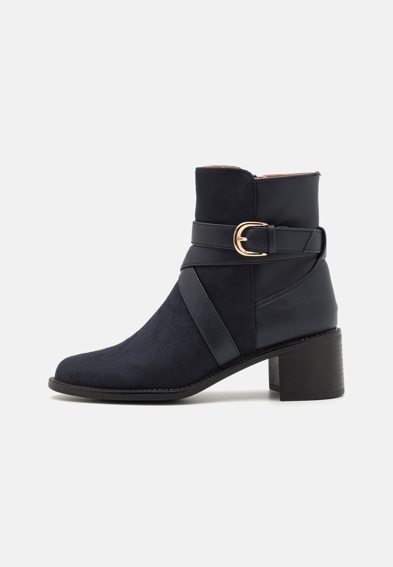 Botte noire à la cheville avec talon bloc, panneaux en daim et cuir, et une sangle décorative avec une boucle dorée autour de la cheville.