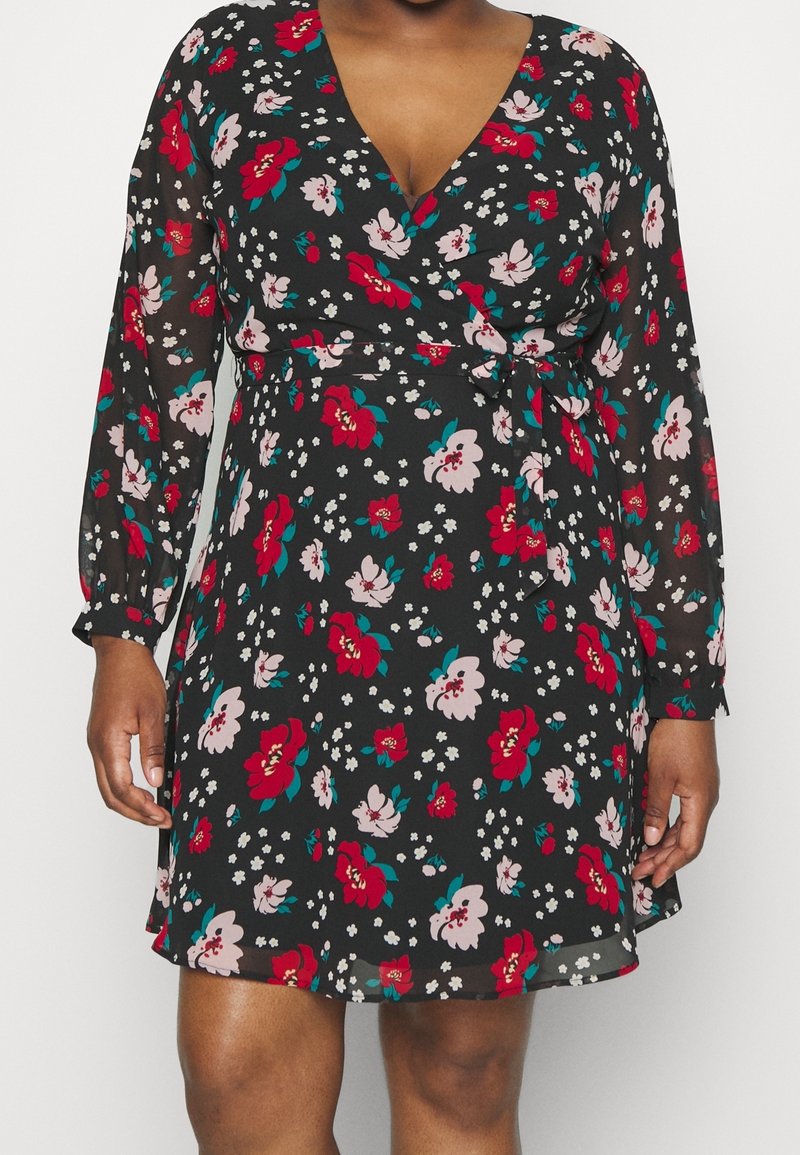 Femme portant une robe portefeuille à manches longues noire avec des motifs floraux rouges, roses et blancs et une ceinture nouée à la taille.