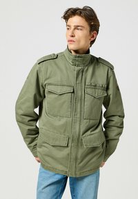 Olivgrön militärjacka i bomull med hög krage, två bröstfickor och två nedre fickor. Slät textur.