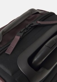 Black travel bag with a textured surface, featuring a grey handle, zip closure, and a maroon accent on the zipper pull.