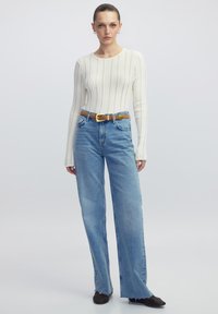 Weißes geripptes Langarmshirt, blaue hochgelegene weit geschnittene Jeans, brauer Gürtel mit goldener Schnalle, schwarze strukturierte Schuhe.