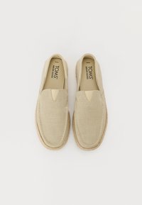 Zapatos slip-on beige con parte superior de lona, que presentan un corte en forma de V en la parte frontal, detalles de costura y una suela texturizada.