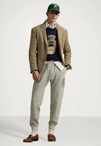 Man i grön keps, beige kavaj, marinblå tröja med björnmotiv, vit skjorta, grå träningsoverallsbyxor och bruna loafers som står mot en enfärgad bakgrund.