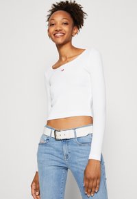 Vit långärmad crop top med rund halsringning och en liten röd logotyp. Kombinerad med ljusblå jeans och ett vitt bälte.