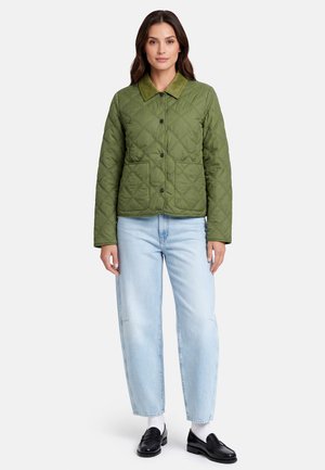 Woman wearing a green quilted jacket with a corduroy collar, light blue jeans, white socks, and black loafers, standing against a white background.