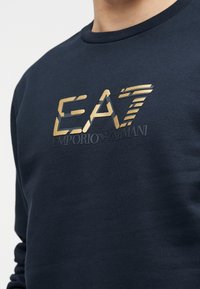 Tumesinine dressipluus pehmest kangast, millel on reljeefne kuldse ja halli värviga logo, mis kannab teksti "EA7 EMPORIO ARMANI" triibuliste aktsentidega.