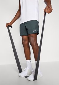 Nike trainingsshorts in donkergroen, gecombineerd met witte sportschoenen en witte kousen. De persoon houdt een grijze weerstandsband vast.