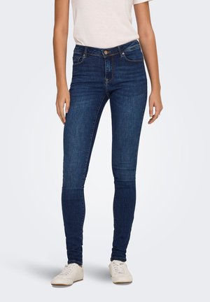 Slanke jeans i mørkeblå denim med mellemhøj talje, for- og baglommer samt minimale syningsdetaljer, parret med hvide sneakers.