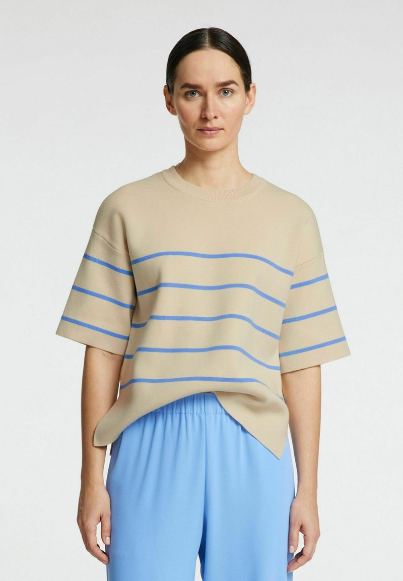 Maglione beige a maniche corte con strisce orizzontali blu, scollatura rotonda e vestibilità comoda. Abbinato a pantaloni larghi blu chiaro.