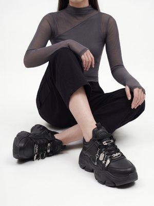 Persona que lleva zapatillas negras voluminosas con colgantes plateados, pantalones negros y una camiseta de manga larga gris translúcida sentada con las piernas cruzadas sobre un suelo blanco.