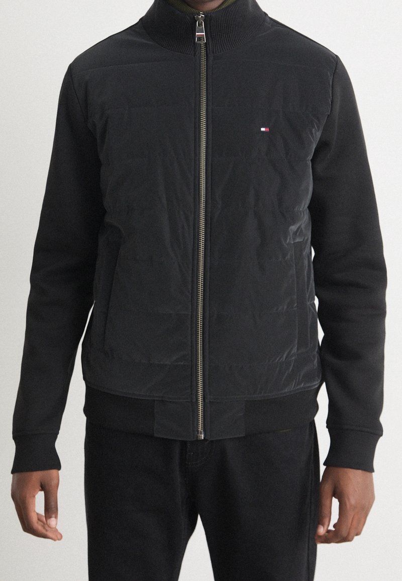 Giubbotto bomber nero con colletto alto, chiusura con zip e polsini a righe. Presenta texture contrastanti e una piccola toppa con logo sul petto.