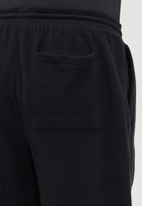Schwarze Hose aus strukturiertem Stoff, mit einer einzelnen aufgesetzten Tasche auf der Rückseite und einem elastischen Bund für zusätzlichen Komfort.