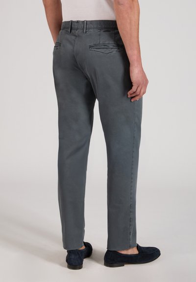Uomo che indossa pantaloni slim grigi e mocassini scuri, mostrato di spalle su uno sfondo chiaro e uniforme.