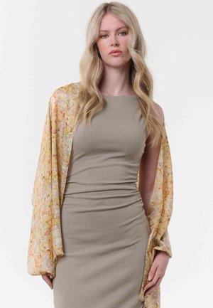 Blonde Frau mit welligem Haar, die ein eng anliegendes, ärmelloses beiges Kleid trägt und einen lockeren, blumigen Schal über die Schultern gelegt hat.