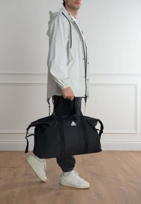 Borsa duffel nera con manici robusti, dotata di una patch con logo, realizzata in materiale durevole, design elegante, scomparti con zip e cinghie laterali.
