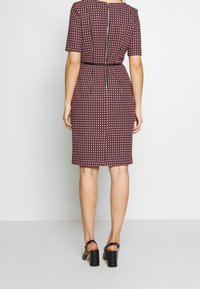 Robe bordeaux à manches courtes avec un motif géométrique, silhouette ajustée et fermeture éclair au dos. Associée à des sandales à talons épais noires.
