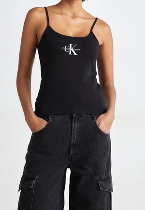 Person trägt ein schwarzes Calvin Klein Jeans Spaghettiträger-Top und schwarze Cargo-Hosen mit Händen in den Taschen vor einem einfarbigen Hintergrund.